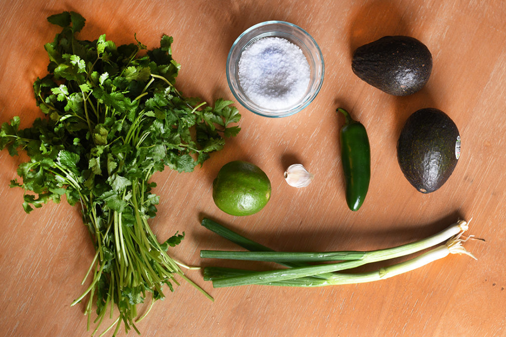 Ingredients for Guacamole: cilantro, green onion, garlic avocado, jalapeño, lime and salt on table